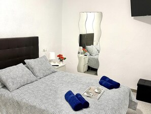 A hotel room with a bed, a bedside table with a lamp and flowers, a mirror, and a tray with towels and a cup. - APARTBEACH OASIS YOUR PARADISE WITH TROPICAL POOL, JACUZZI (RIUDOMS)