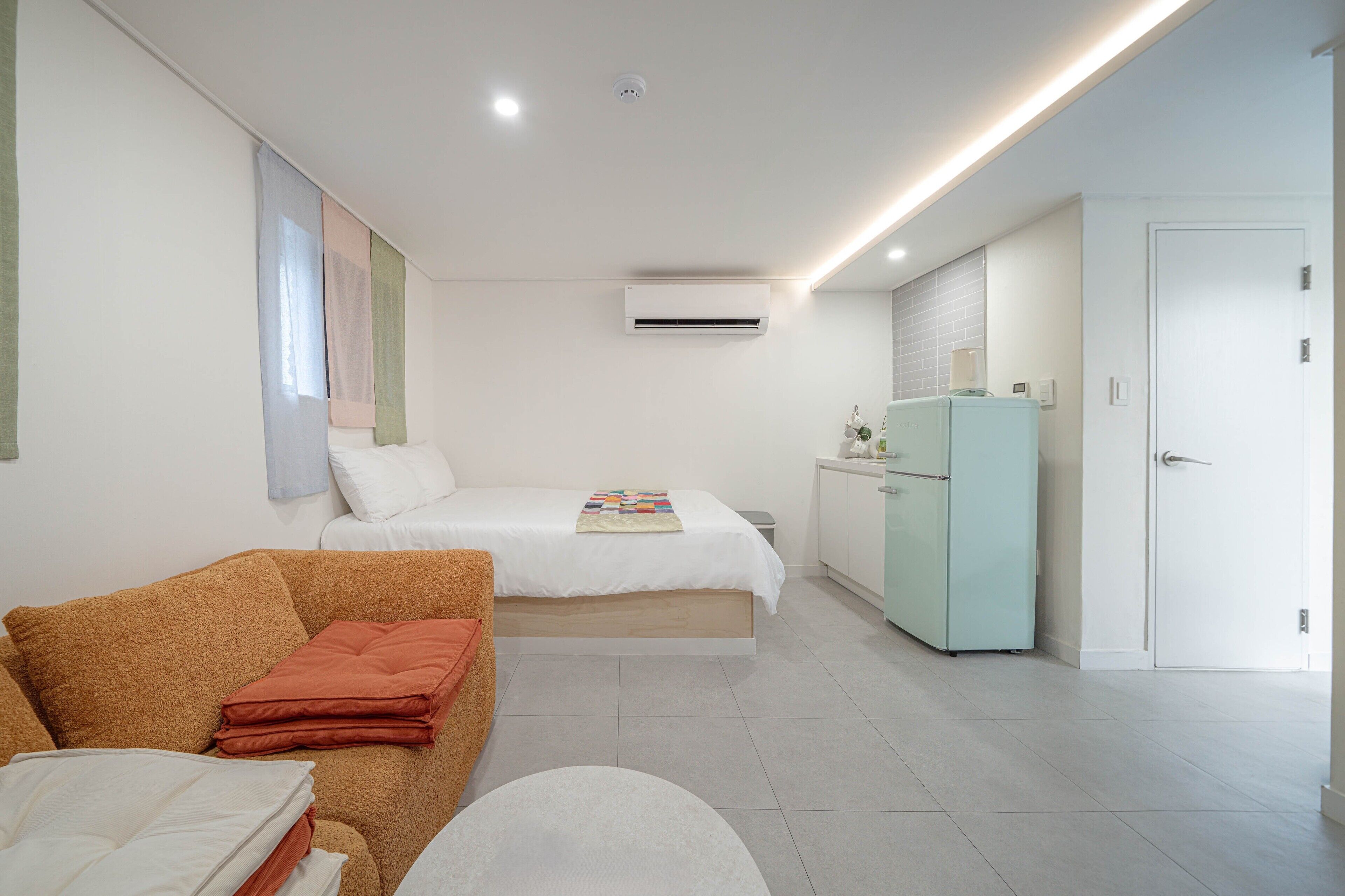 A modern hotel room with a bed, a sofa, a small table, an air conditioning unit, a refrigerator, and a door.