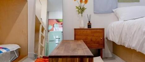 A bedroom with a bed, a wooden table, a nightstand, a mirror, and a window.