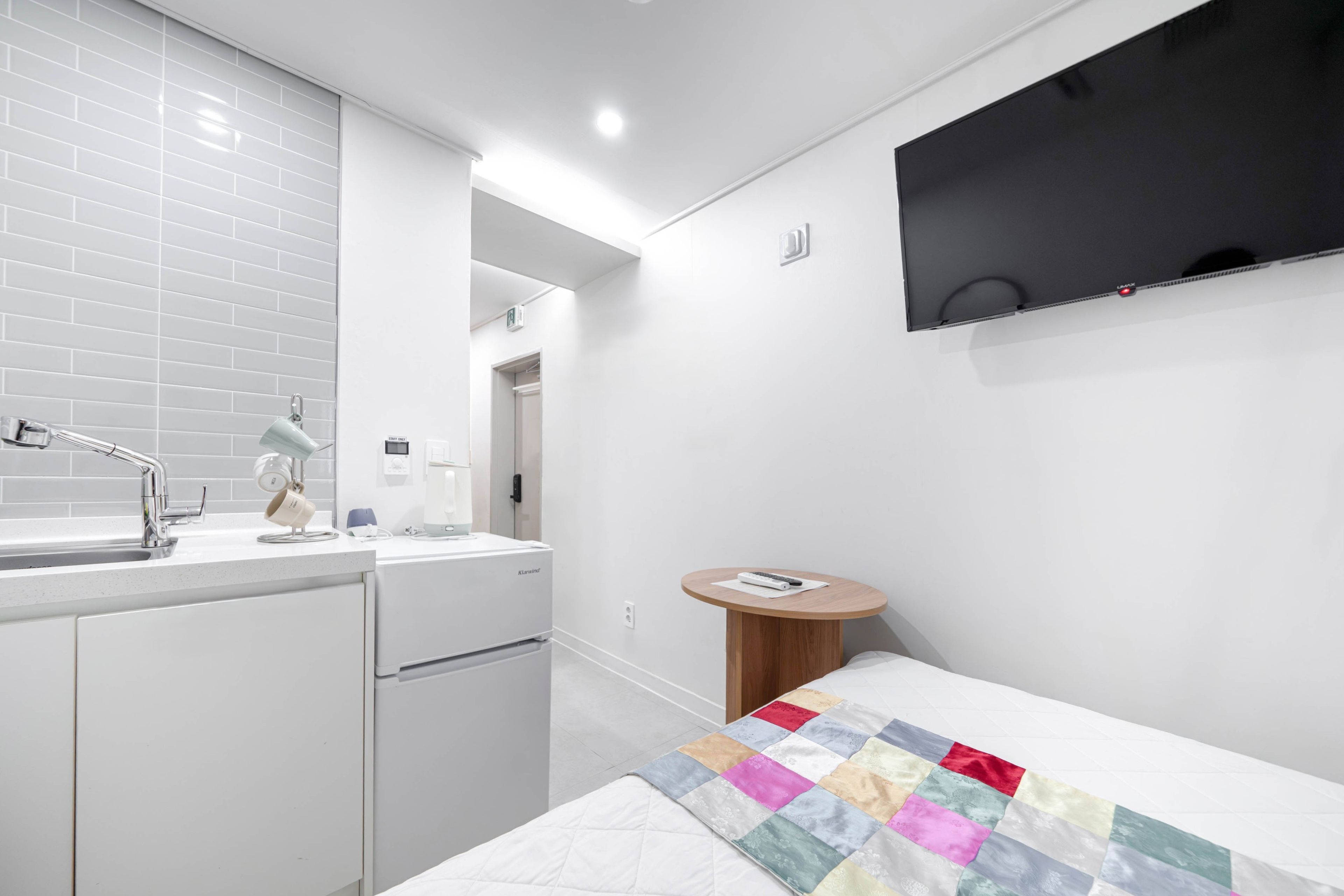 Une chambre d’hôtel moderne équipée d’une kitchenette, d’un téléviseur à écran plat fixé au mur, d’une petite table ronde en bois et d’une couverture colorée sur le lit.
