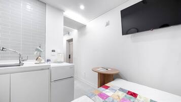 Une chambre d’hôtel moderne équipée d’une kitchenette, d’un téléviseur à écran plat fixé au mur, d’une petite table ronde en bois et d’une couverture colorée sur le lit.