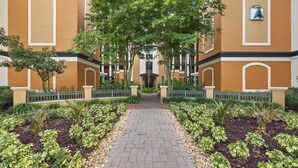 Uma trilha leva a um edifício com fachada simétrica, entrada central e um jardim circundante com diversas plantas.