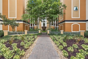 A pathway leading to a building with a symmetrical facade, a central entrance, and a surrounding garden with various plants. - 2BR Condo 7 mins to Disney | FREE Resort Access (Orlando)