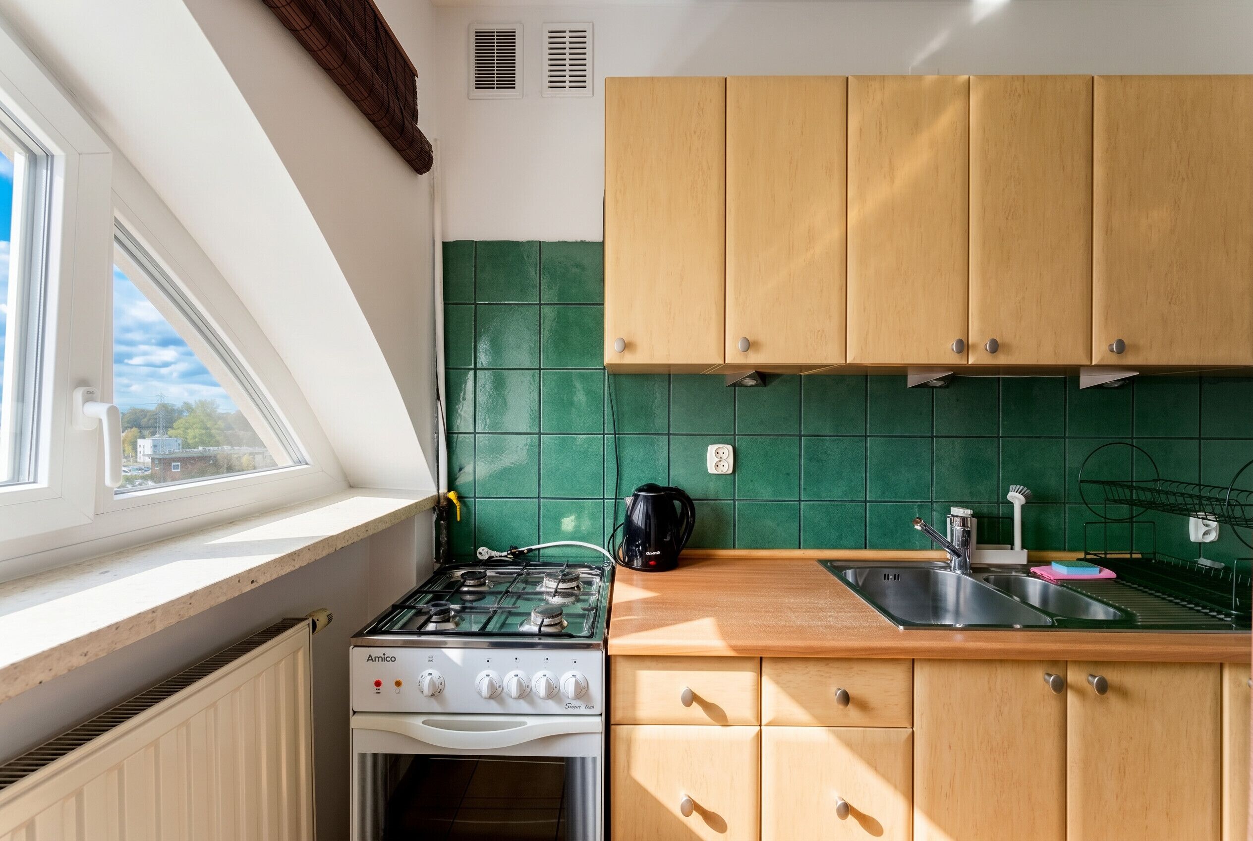 Een keuken met een gasfornuis, oven, houten kastjes, een raam en een groene betegelde achterwand.