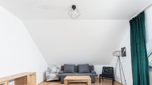 Una habitación moderna estilo loft con piso de madera, un sofá gris, una mesa de centro de madera, un sillón estampado, una silla negra, una lámpara de pie y un ventanal grande con cortina.