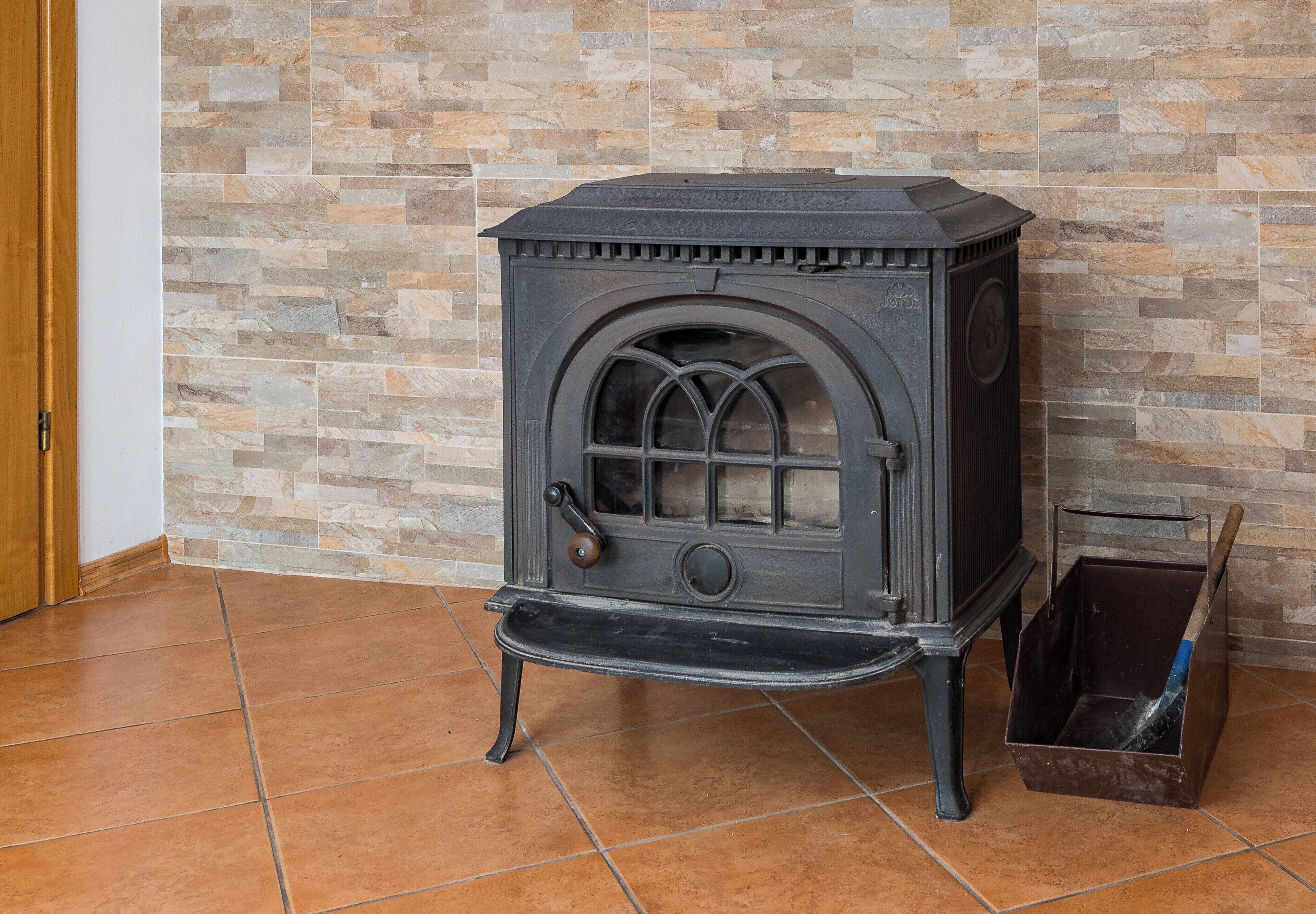 Un horno a leña negro con puerta de vidrio, un balde de metal y una pared con diseño de piedra al fondo.