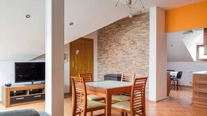 A dining area with a glass table, wooden chairs, a television on a wooden stand, and a stone wall.