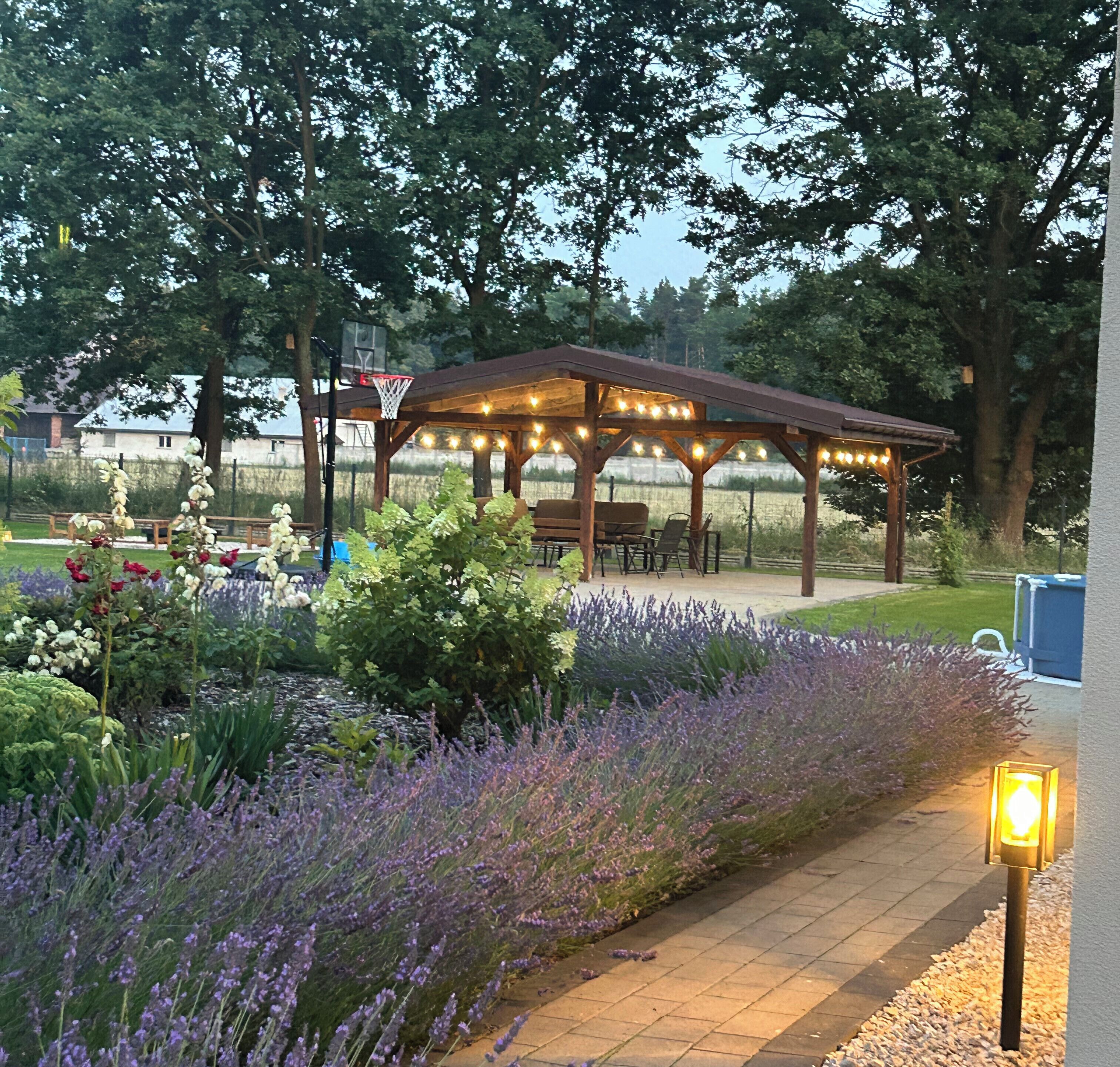 Un espace extérieur aménagé avec des sièges, protégé par un auvent et illuminé de guirlandes lumineuses, entouré d’un jardin où poussent de la lavande et d’autres plantes.