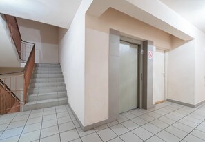 Eine Treppe mit hölzernen Geländern führt zu einer Zwischenebene, eine geschlossene Aufzugstür und ein Rauchverbotsschild an der Wand.