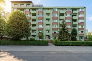 Ein mehrgeschossiges Gebäude mit grüner Fassade, mehreren Balkonen und einem gepflasterten Eingangsbereich.