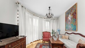 A living room with a TV, a red chair, a sofa, a chandelier, and a colorful painting on the wall.