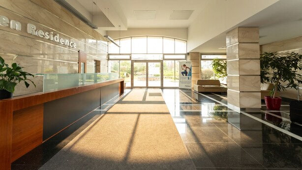A modern hotel lobby with a reception desk, a glass partition, and a large glass door leading outside.