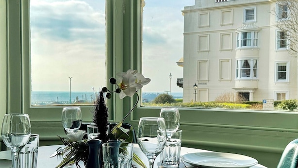 Une table à manger dressée avec des verres et un vase de fleurs, offrant une vue sur la côte et un grand bâtiment.