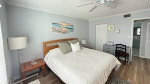 Ein Schlafzimmer mit einem Bett, einem Schreibtisch, einem Stuhl, einem Deckenventilator, einem Nachttisch, einer Lampe, einem Gemälde und einem Kleiderschrank.