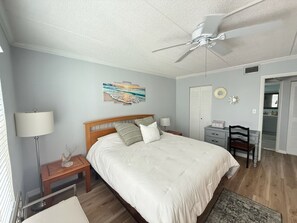 Ein Schlafzimmer mit einem Bett, einem Schreibtisch, einem Stuhl, einem Deckenventilator, einem Nachttisch, einer Lampe, einem Gemälde und einem Kleiderschrank.