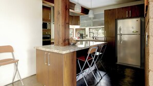 A kitchen with wooden cabinets, a stainless steel refrigerator, a marble countertop, and a bar area with chairs.
