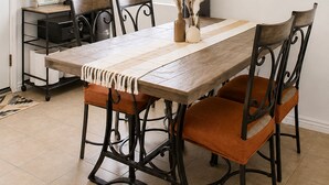 Un comedor con una mesa de madera, cuatro sillas, una lámpara, una ventana con cortinas y una obra de arte colgada en la pared.