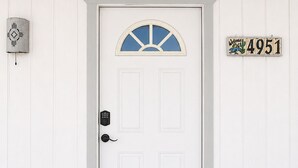 Una puerta blanca con un ventanal de cristal azul, un timbre, un cartel de bienvenida y una placa de matrícula.
