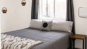 Un dormitorio con cama, ventilador de techo, ventana con cortinas y una mesita de noche pequeña.