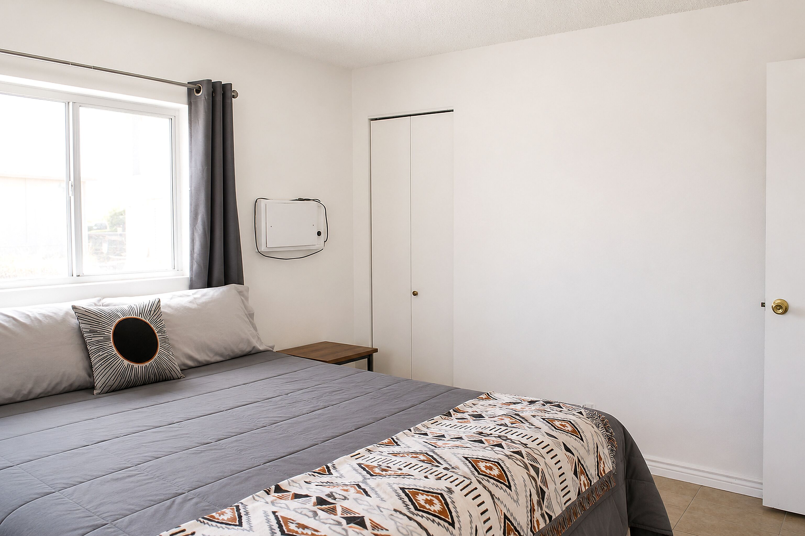 A bedroom with a bed, a window with curtains, a bedside table, and a wall-mounted heater.