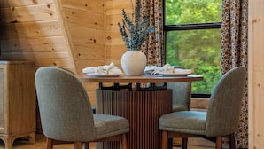 A wooden cabin interior with a dining table, two chairs, a vase with plants, and a window with curtains. - Luxury A-Frame w/ Hot Tub (Sevierville)