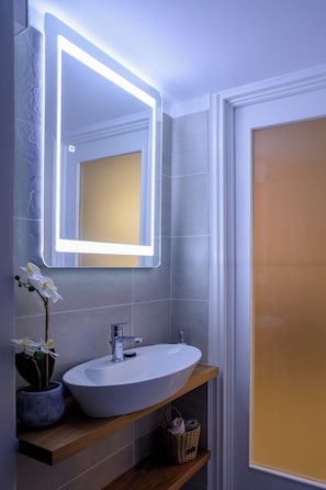 A bathroom with a white sink, a mirror with illuminated edges, a potted orchid, and a wooden shelf with a basket. - Athenian Residential Apartments (Athens)