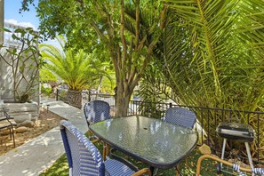 Une terrasse avec une table en verre et des chaises bleues, entourée d’une végétation luxuriante et d’une clôture en métal.