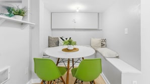 Moderni huone, jonka seinät ovat valkoiset. Huoneessa on pyöreä pöytä, kaksi vihreää tuolia, ruukkukasvi ja tyynyillä varustettu penkki.