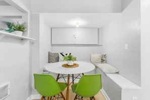Una habitación moderna con paredes blancas, una mesa redonda, dos sillas verdes, una planta en maceta y un banco con cojines.