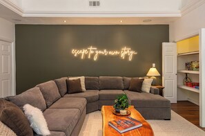A modern living room with a sectional sofa, a coffee table with a book, and a neon sign on the wall. - The Fable House | Sequoia Riverfront Modern Design (Three Rivers)