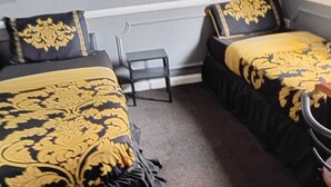 Une chambre avec deux lits, chacun recouvert d’une couvre-lit à motifs noirs et dorés, une petite chaise noire et un mur gris avec une prise électrique blanche.