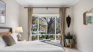 A bedroom with a large window, a bed, a bedside lamp, a plant, and a mirror. - Salt & Stay - Laid-Back Comfort by the Beach (Encounter Bay)