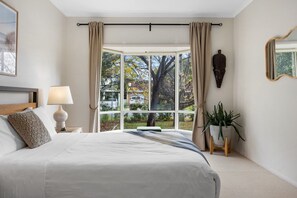 A bedroom with a large window, a bed, a bedside lamp, a plant, and a mirror. - Salt & Stay - Laid-Back Comfort by the Beach (Encounter Bay)