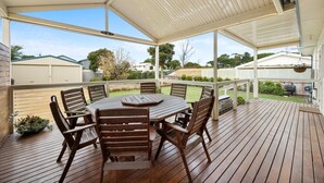 A covered patio with a wooden table and chairs, a view of the backyard, and a white fence. - Salt & Stay - Laid-Back Comfort by the Beach (Encounter Bay)