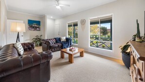 A spacious living room with a leather sofa, a blue armchair, a wooden coffee table, and large windows offering a view of the outdoors. - Salt & Stay - Laid-Back Comfort by the Beach (Encounter Bay)