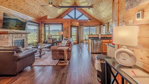 Ein rustikales Holzhaus-Interieur mit Kamin, Ledersofa, Essbereich, Küche und großen Fenstern mit Blick auf die Berge.
