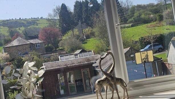 Une vue par la fenêtre sur un paysage rural, avec des maisons, une figurine de cerf et un vase rempli de fleurs blanches.