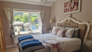 Une chambre avec un grand lit, une fenêtre avec des rideaux, une table de chevet avec une lampe et un tableau floral accroché au mur.