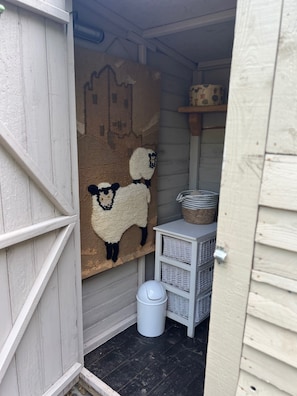 Een kleine, afgesloten ruimte met een witte deur, een wanddecoratie van geweven wol met schapen, een rieten mand en een witte prullenbak.