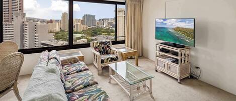 Habitación de hotel con un ventanal grande, un sofá, una mesa de centro de cristal, un televisor y una mesita con mueble para televisión.