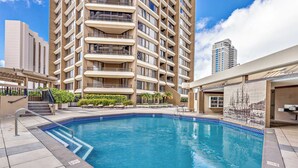 A swimming pool with steps, a building with balconies, and a clear sky. - 28th Floor Ala Moana Suite | City + Mountain Views (Honolulu)
