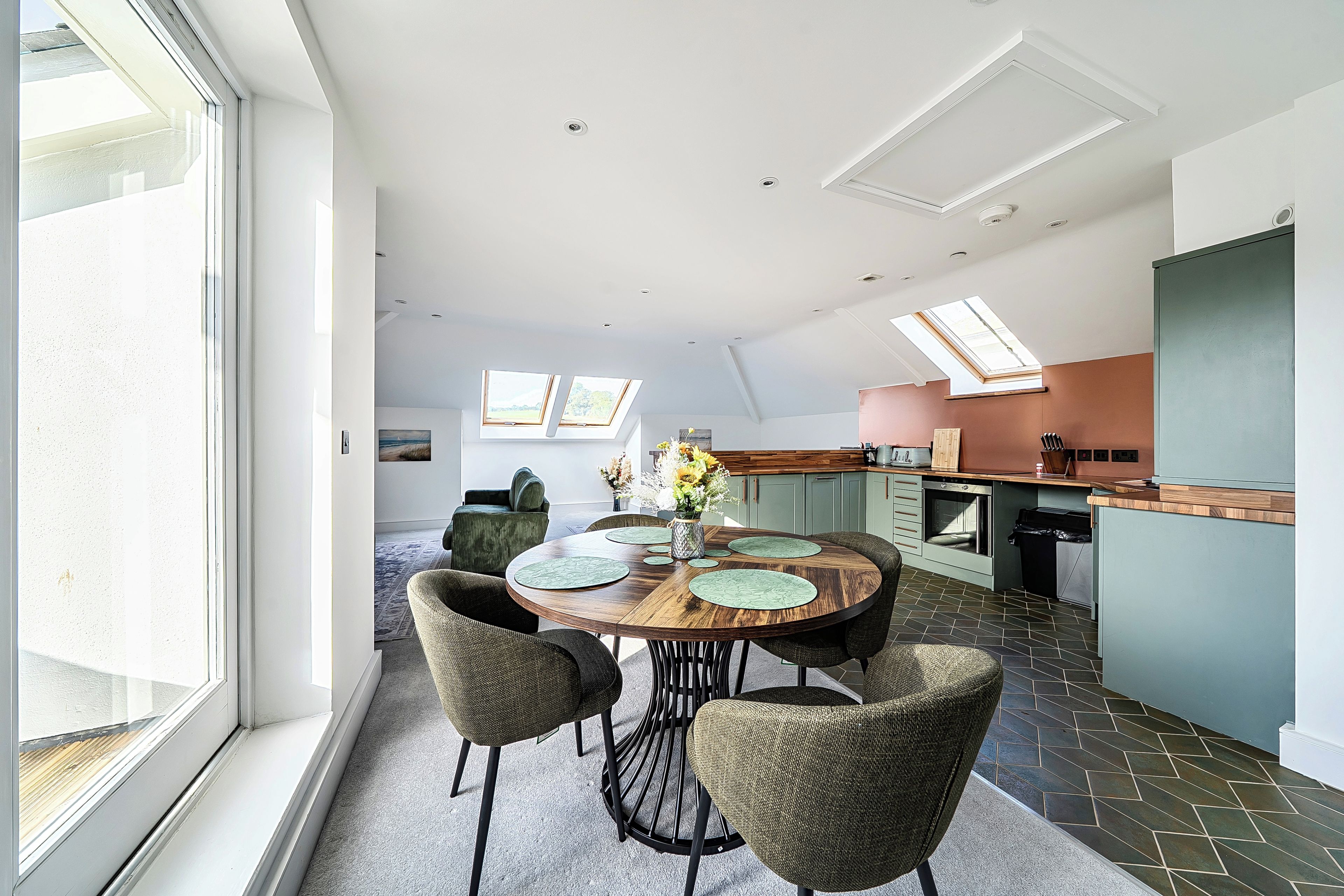 A modern kitchen with a dining area, featuring a round wooden table with chairs, a built-in oven, and skylights.