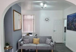Een woonkamer met een grijze bank, een rond bijzettafeltje, een spiegel, een zonachtige decoratie aan de muur, en een deur die leidt naar een andere kamer.