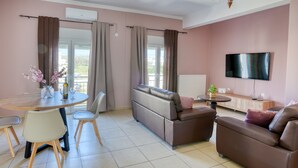 Ein Wohnzimmer mit einer Couch, einem Fernseher, einem Esstisch mit Stühlen und einem Balkon mit Vorhängen.