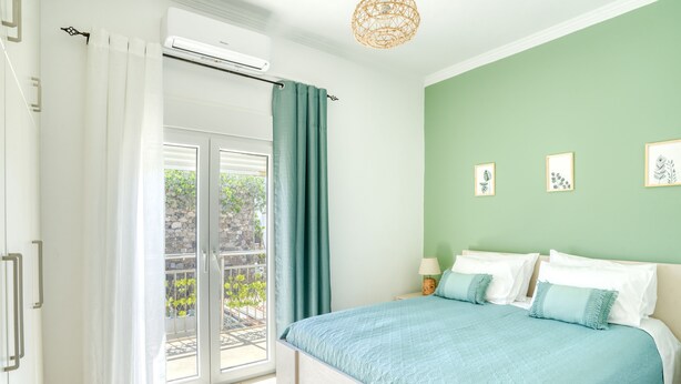 Ein Schlafzimmer mit Bett, einem Fenster mit Vorhängen, einem Balkon und einer Wand mit gerahmten Bildern.