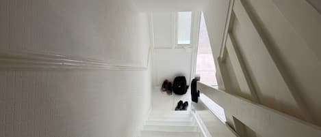 Un escalier blanc avec une rampe, menant à un palier où sont posées des chaussures et un sac à dos.