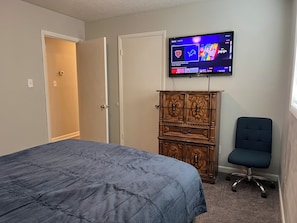 Un dormitorio con una cama, un televisor montado en la pared, un aparador de madera y una silla azul.
