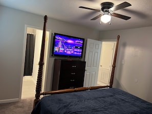 Un dormitorio con cama con dosel, ventilador de techo, televisor sobre un tocador y puerta que da a otra habitación.