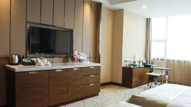 Ein Hotelzimmer mit großem Flachbildfernseher, einer Minibar, einem Schreibtisch mit Stuhl, einem Bett und einem großen Fenster.