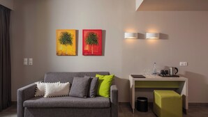 A modern living room with a grey sofa, two wall-mounted paintings, a white desk with a lamp, and a small green ottoman. - Unique Blue Resort (Hersonissos)